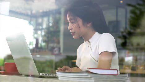 portrait from a window of an asian teenage girl working on a laptop in a cozy coffee shop on a bright sunny day, enjoying a relaxed atmosphere with warm natural light streaming into the café