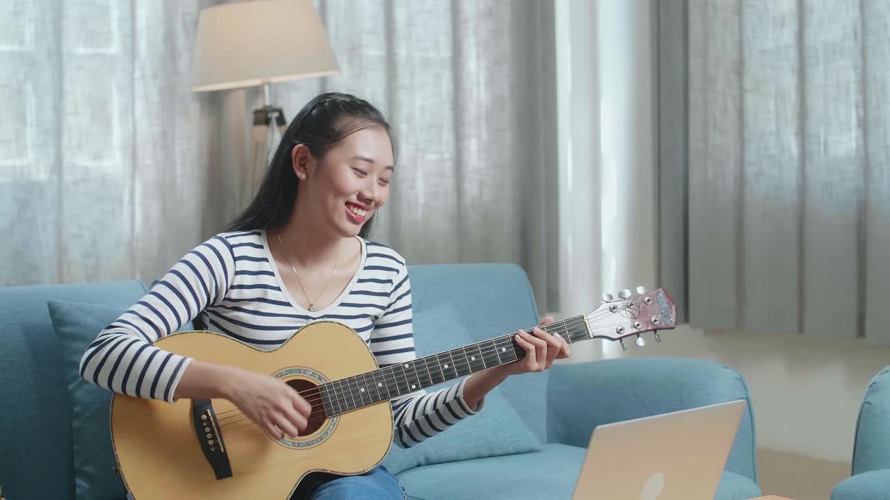 노트북을 들고 있는 미소 짓는 아시아 여성이 집에서 기타를 연주하는 것을 즐기고 있습니다.