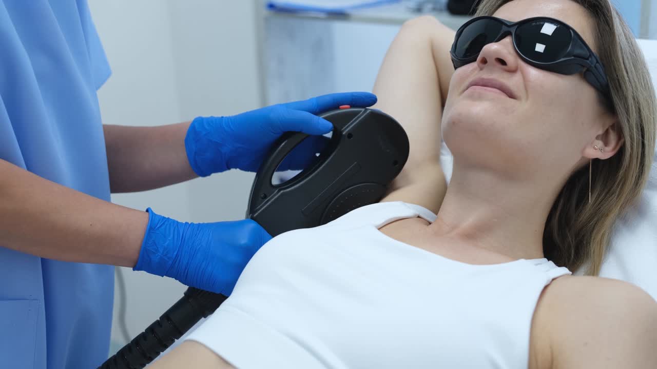 보호 안경을 입은 여성 고객이 미용실에서 팔꿈치 레이저 제거를 받고 있습니다.