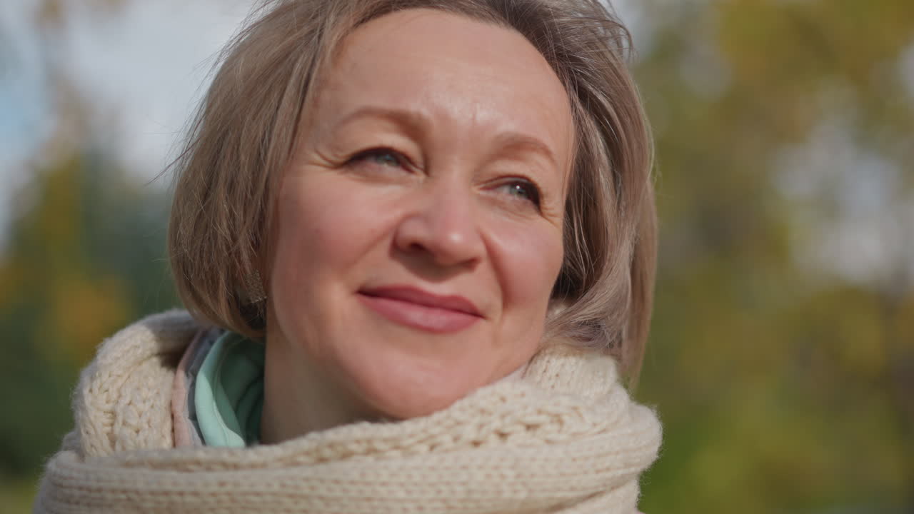 middle age woman with serene smile gazing upward while gentle breeze lifts hair and scarf, soft sunlight highlighting peaceful emotion with blurred trees and sky forming tranquil background