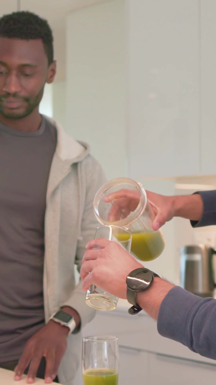 Video vertical: Hombre con sudadera revisando el teléfono en la cocina mientras un amigo vierte jugo en un vaso.
