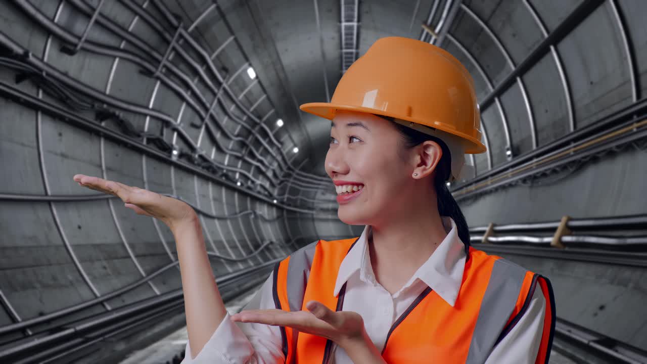 Close Up Of Asian Female Engineer With Safety Helmet Smiling And Pointing To Side In Underground Subway Tunnel
