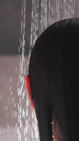 woman turns around head under flow of water. lady moves around head with brunette hair under spray of water in shower with greyness of tiled wall