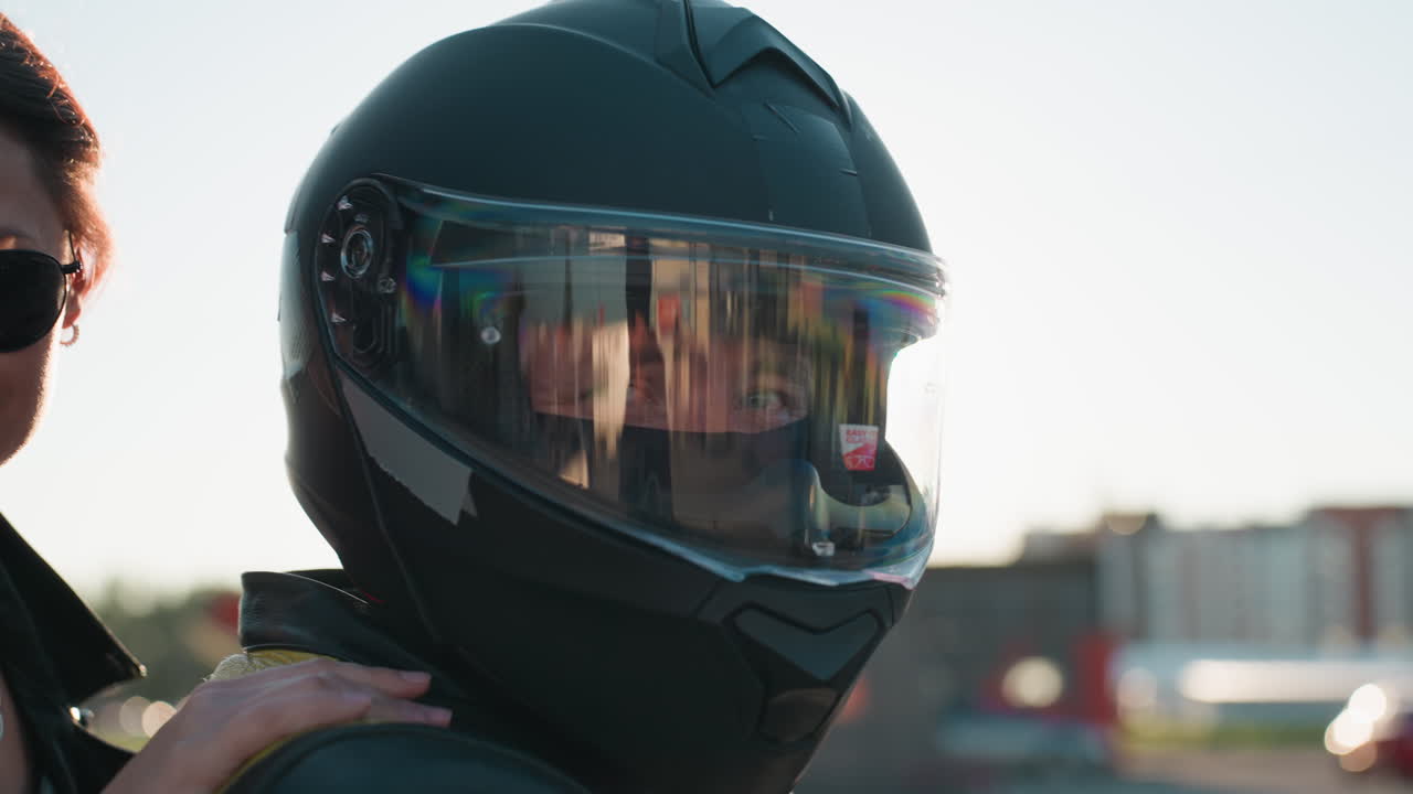 Rider wearing helmet sits on motorcycle with sunlight creating glow effect on visor while passenger behind rests hand on shoulder, both looking calm in stylish black jackets under clear bright sky