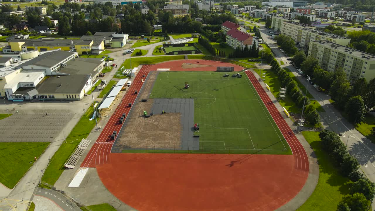 Aerial drone footage ascending and flying higher above a large modern green and red colored soccer field that is being built or renovated by workers that have machines, summer sunny day, urban area