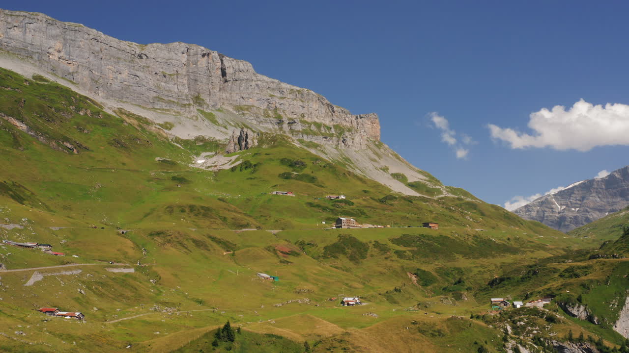 Alpine hamlet beneath towering mountain cliffs. Mountain landscape with lush green meadows under a vivid blue sky. Pan right. Switzerland