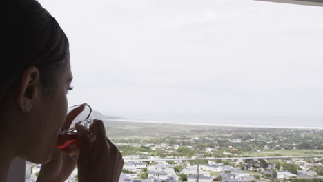 thoughtful biracial woman looking through window drinking tea at home, copy space, slow motion