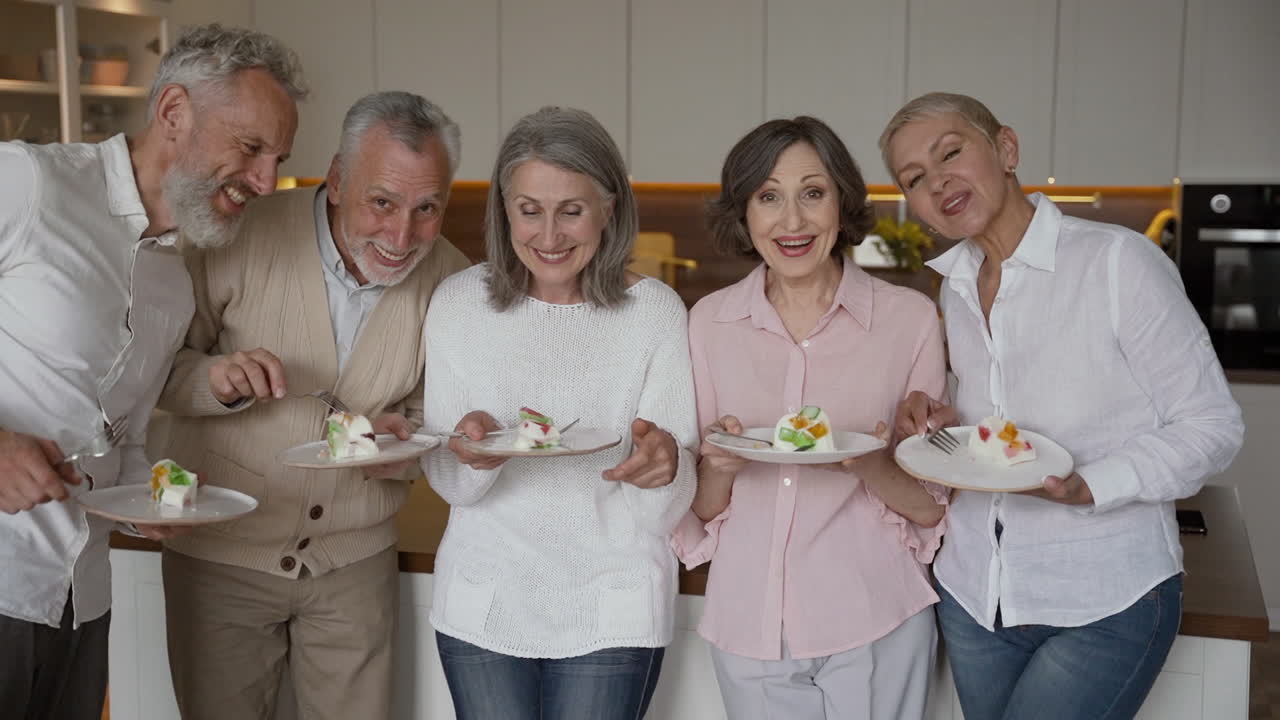 Gruppe glade seniorvenner har det gøy med å riste tallerkener med flan, mens de står og ser mot kameraet på et kjøkken