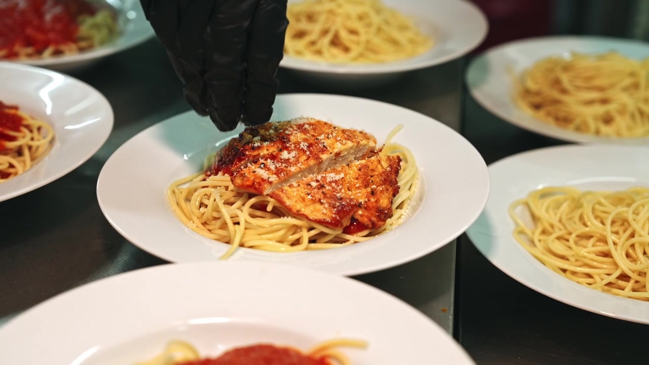Ripresa ravvicinata in tracking della mano di uno chef che sparge finemente parmigiano grattugiato ed erbe aromatiche essiccate su pollo grigliato a fette adagiato su piatti di spaghetti al pomodoro