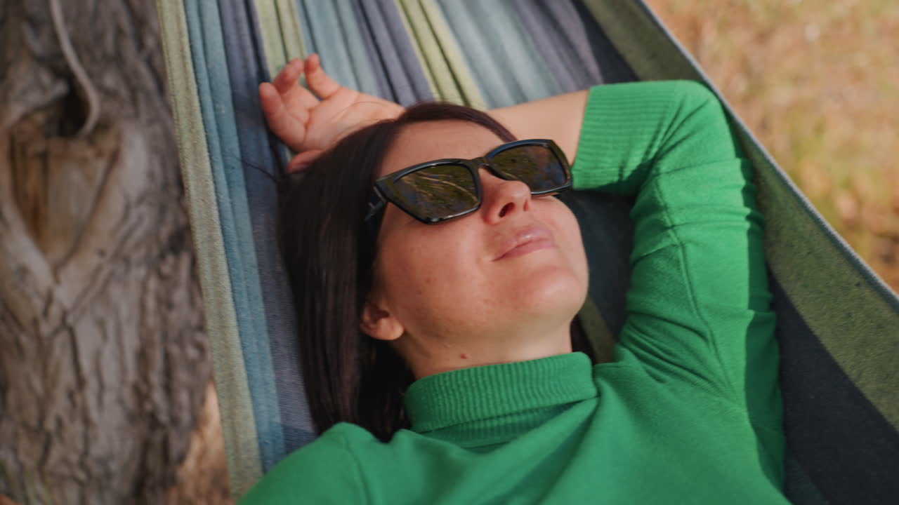 Donna reclinata in amaca in una scena di campeggio sotto un albero, maglione verde e occhiali da sole, foglie autunnali a terra, soffice luce solare sul viso, occhi chiusi, riposo solitario e pacifico, umore calmo e meditativo, respiro lento