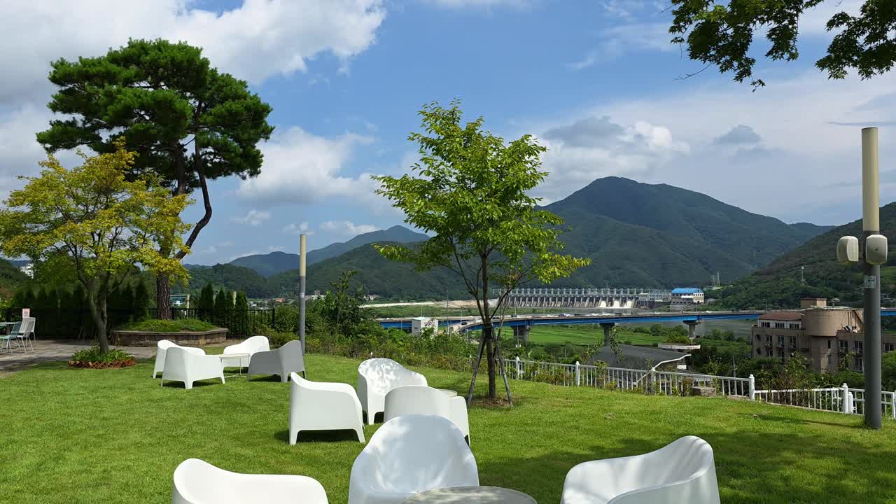 Relaxing walking point of view shot moves through a cozy terraced cafe garden, revealing a stunning panorama of the Bukhangang River, New Cheongpyeong Bridge and Dam nestled in Gapyeong mountains