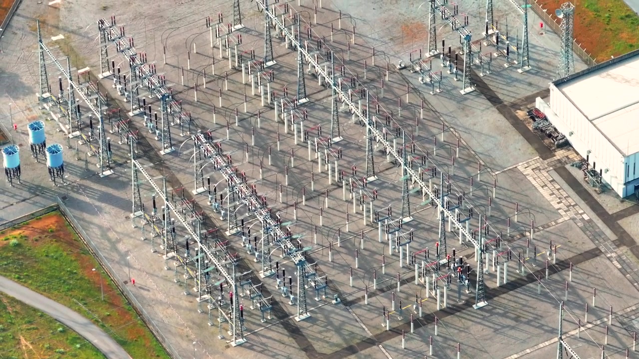Luftaufnahmen eines Umspannwerks. Hochspannungsausrüstung und Übertragungsleitungen sind sichtbar. Das Layout umfasst verschiedene Strukturen und Wege in einer Umgebung der elektrischen Infrastruktur.