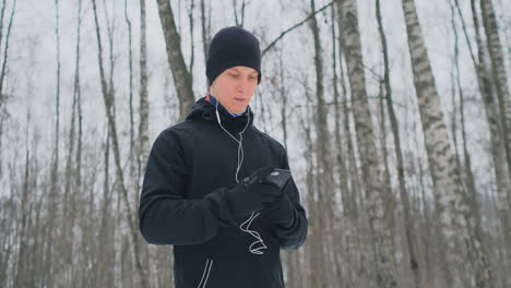 Un-Joven-Con-Un-Martillo-Negro-En-Invierno-Se-Prepara-Para-Correr-En-El-Parque,-Incluye-Una-Aplicación-Para-Correr-Y-Corre-Con-Un-Asistente-De-Voz-Por-El-Bosque-Haciendo-Negocios-Y-Llevando-Un-Estilo-De-Vida-Saludable.