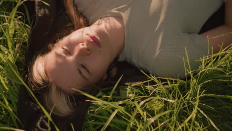 woman lying peacefully on cloth spread over grassy field, surrounded by tall green blades of grass and warm sunlight casting gentle shadows on her face