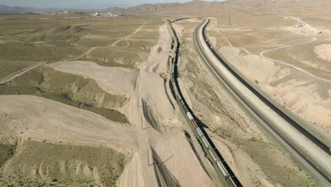 Long-Cargo-Train-Traveling-Through-Nevada-State-Next-To-Vehicles-Driving-On-Asphalt-Road-In-USA