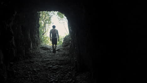 hiker with an orange backpack in summer clothes walking throug a dark tunnel carved in the rock towards the light where are visible green trees