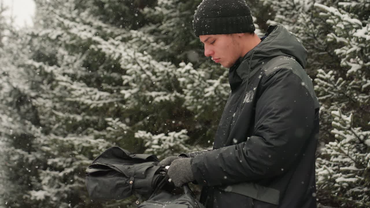 Caucasian man snowy pine forest adjusting gloves, checking backpack pocket while snowflakes fall, black jacket and beanie, soft overcast light, contemplative expression, close profile, serene outdoor