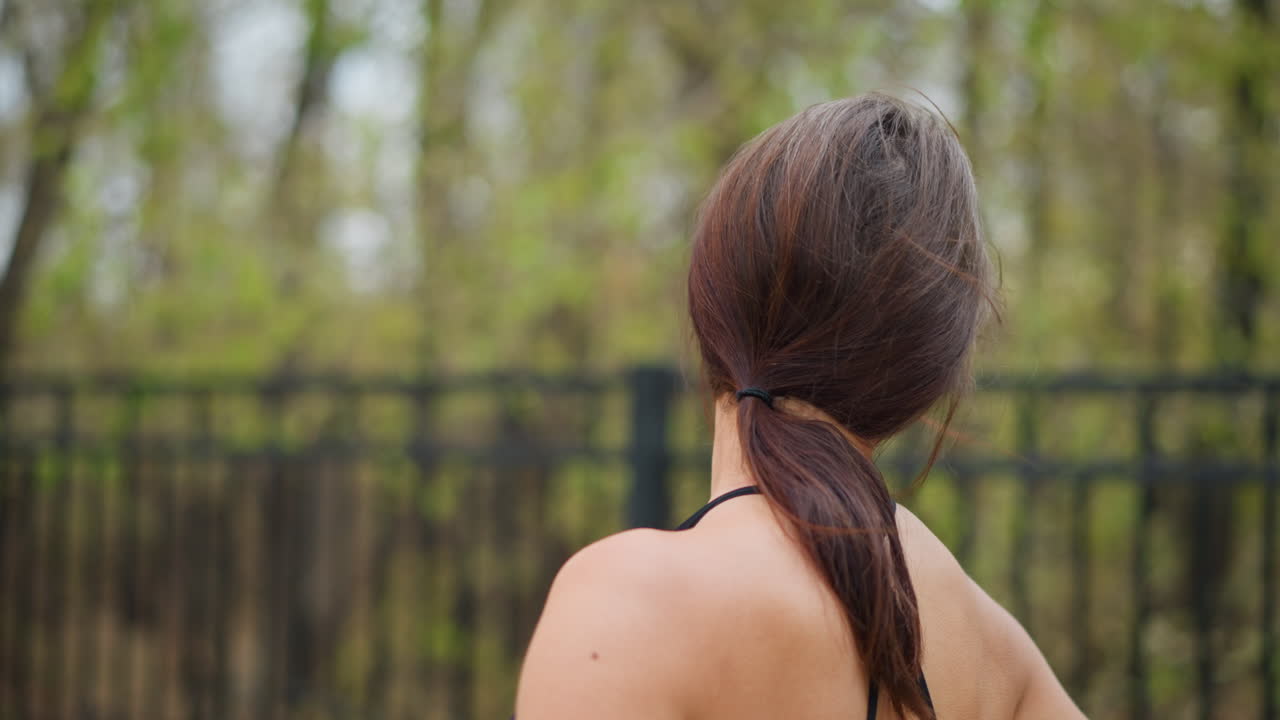 Portrait back view of young woman stretching neck gently outdoors with blurred iron fence and trees in background, promoting relaxation and flexibility in natural setting