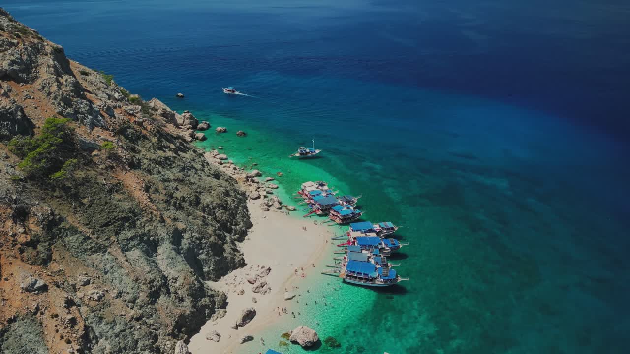 Türkiye'deki Suluada Adası'nın hava drone görüntüsü; kayalık uçurumların altında kumsalı ve kristal berraklığında turkuaz Akdeniz sularında demirlemiş çok sayıda tur teknesiyle.