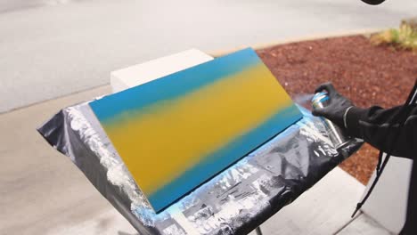 man with black gloves holding spray paint can spraying it across a wooden canvas board with blue yellow wih street in background