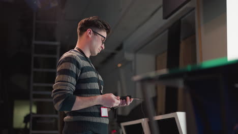 man wearing striped sweater and glasses adjusts wireless audio transmitter on video production set, with cable and equipment on white table, indoor studio with ladder and background lighting