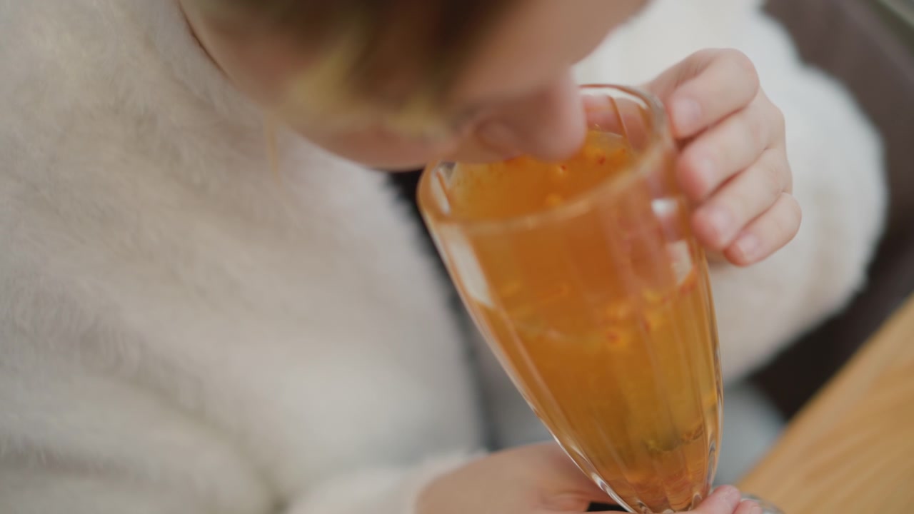 Vista ravvicinata dall'alto mostra una mano adulta che culla un alto bicchiere trasparente pieno di una bevanda arancione traslucida, inclinato verso la bocca con una presa salda, increspature visibili, luce interna calda, riflessi tenui