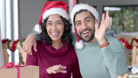 happy biracial couple wearing santa claus hats having christmas video call at home, in slow motion