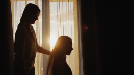 a woman combs her daughter's long hair. sitting by the window at sunset