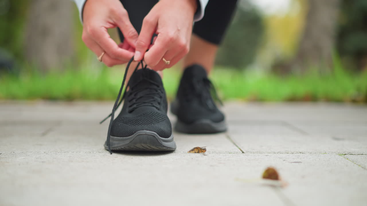 Close-up of lady tying her black sneakers shoelace with focus on hands, showcasing rings, preparing for exercise, outdoor workout in park on sunny day, wearing athletic gear