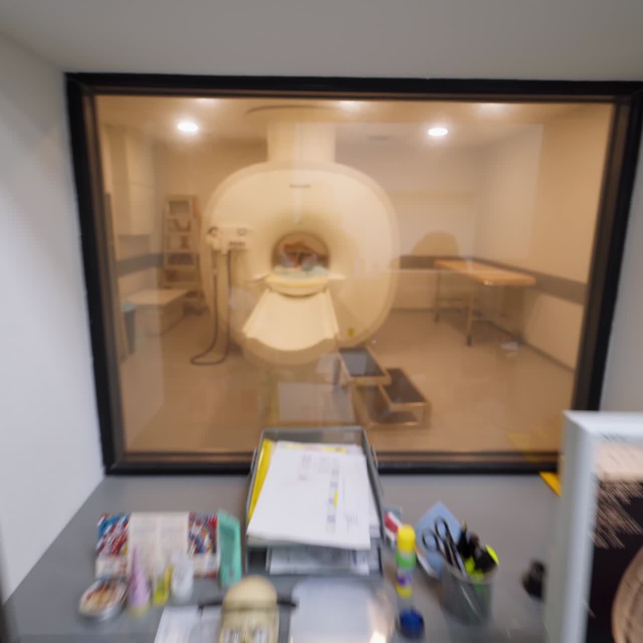 Female laboratory worker enters the room and goes to her seat. Camera follows the medic and shows the magnetic resonance scanner through a little window
