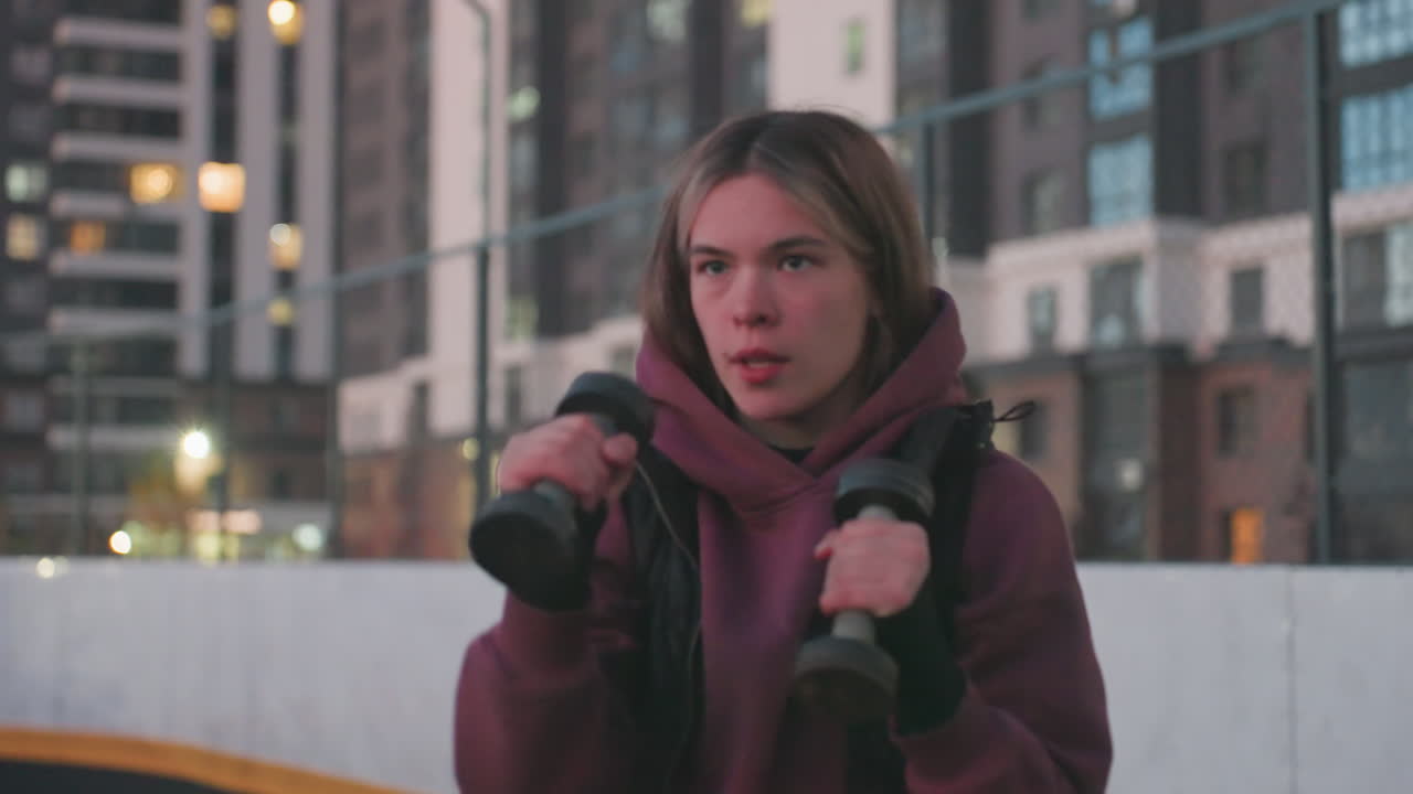 Medium shot of training partner boxing dumbbells punching air mid workout on black asphalt court near white barrier topped with chain link fence under urban dusk sky wearing maroon hoodie