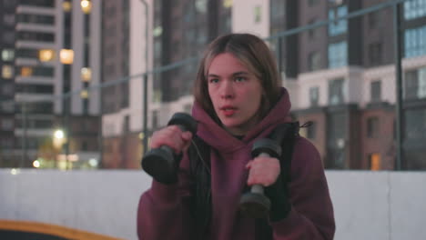 medium shot of training partner boxing dumbbells punching air mid workout on black asphalt court near white barrier topped with chain link fence under urban dusk sky wearing maroon hoodie