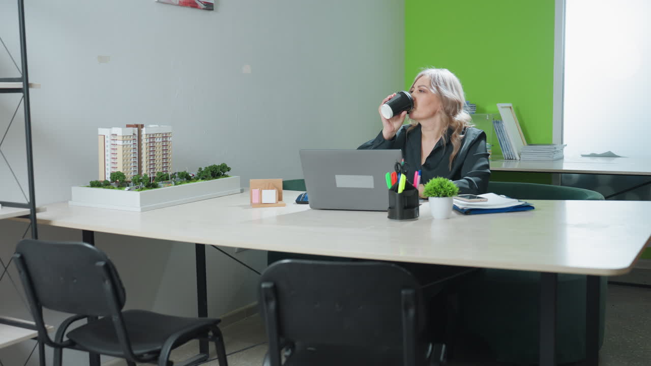 Sales woman seated at office desk working on laptop pauses to take latte before continuing, surrounded by model structure, organized desk items