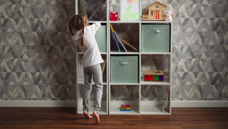 little girl in pajamas takes toys off box on rack at home