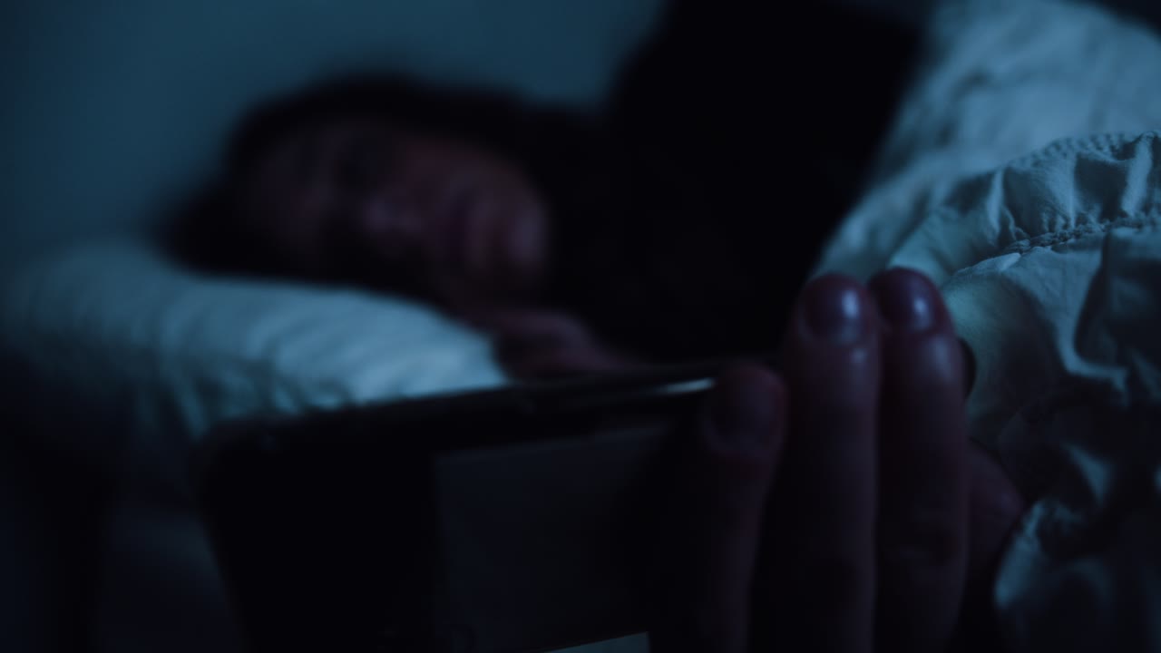 Smartphone utilizzato a letto, con messa a fuoco che passa dal telefono a una donna stanca.