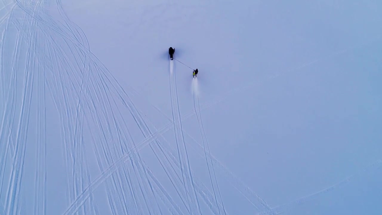 Vista spettacolare di una motoslitta che traina uno sciatore con pantaloni gialli su una montagna innevata con tracce di motoslitta, aerea