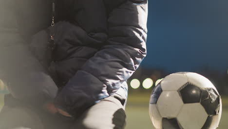 a man sits alone outdoors at night in an empty stadium, dressed in a hooded jacket with his hands clasped between his legs a ball is close to him, with blurred views of a building and dim light