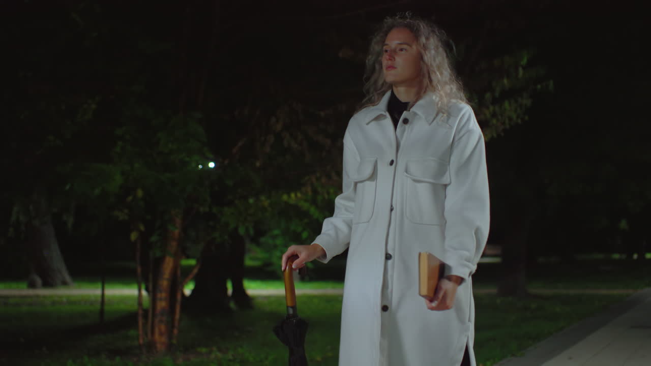 Calm lady in white coat walking alone through open park at night holding umbrella and book, approaching wooden bench surrounded by dark trees and soft ambient lighting