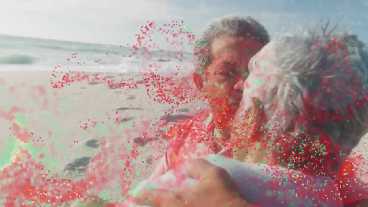 Casal idoso parando na orla, abraçando-se, beijando-se e mostrando a aliança de casamento, com partículas em movimento.