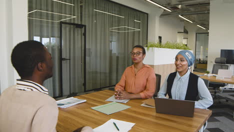 an woman and a muslim woman co workers interview a young man sitting at a table in the office 1