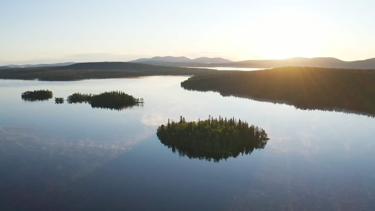 Aerial view high above a blue foggy lake during sunrise showing small islands and mountains in the background as sun is giving golden hour light to the landscape filmed in Lapland Finland