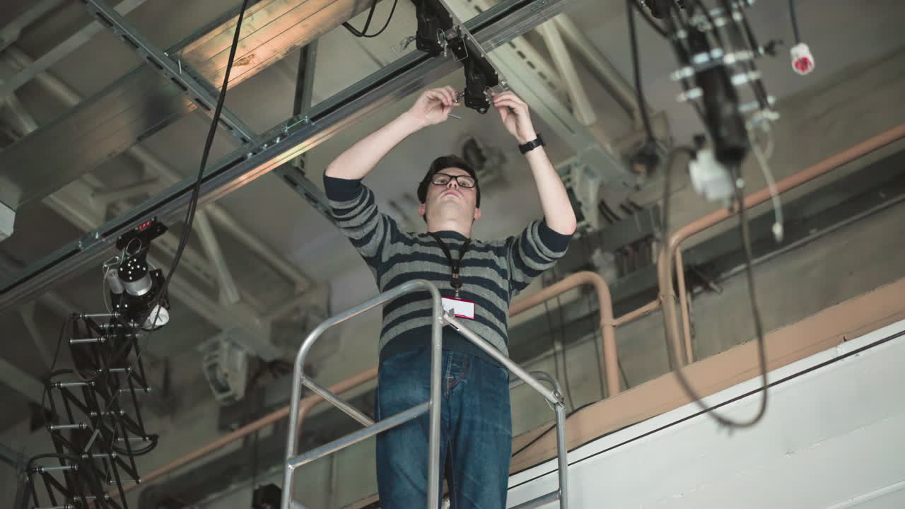 Person working on elevated platform, adjusting camera or lighting equipment mounted on ceiling, wearing sweater and glasses, industrial setting, cables visible, maintenance or installation task