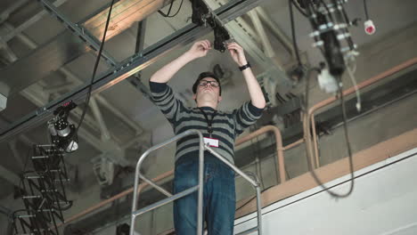 person working on elevated platform, adjusting camera or lighting equipment mounted on ceiling, wearing sweater and glasses, industrial setting, cables visible, maintenance or installation task