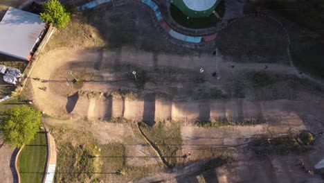 Toma-Aérea-De-Arriba-Hacia-Abajo-De-Un-Ciclista-De-Bmx-Montando-Y-Practicando-En-Una-Pista-De-Carreras-Sucia-Y-Arenosa-Durante-La-Puesta-De-Sol-En-Buenos-Aires,-Argentina