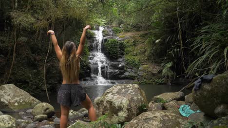 vista trasera de una mujer morena levantando las manos en el aire mientras se enfrenta a una impresionante cascada - retiro de la selva tropical de o&#39;reilly - costa dorada, australia - plano medio