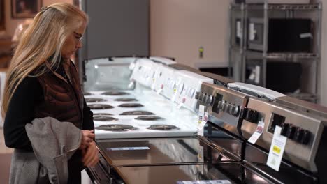 mujer rubia bastante madura girando las perillas de una estufa en una tienda de electrodomésticos de cocina