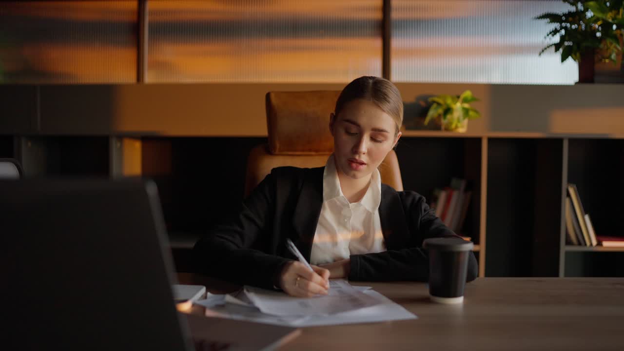 A confident blonde businesswoman in a black jacket and business uniform sits at a table and writes her ideas on important sheets of paper in front of a laptop in a sunny office