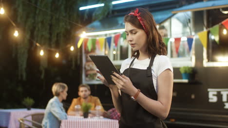 Una-Joven-Y-Bonita-Camarera-En-Delantal-Usando-Un-Dispositivo-De-Tableta-En-El-Parque-En-La-Pista-De-Comida-Al-Aire-Libre