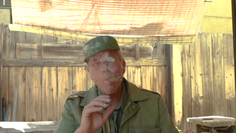 portrait of a mature male in his 60s smoking a cigar, sitting outdoors under a shade cloth