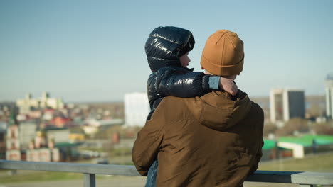 a father carries his son while the child playfully covers his father's ear as they stand by an iron railing, overlooking a bridge with a blurred cityscape and cars passing by